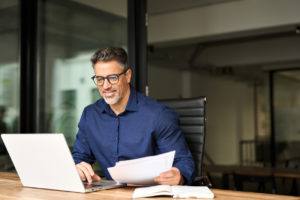 Happy busy older professional businessman entrepreneur using laptop holding bill corporate document in office. 50 years old man executive manager or business owner reading paper working on computer